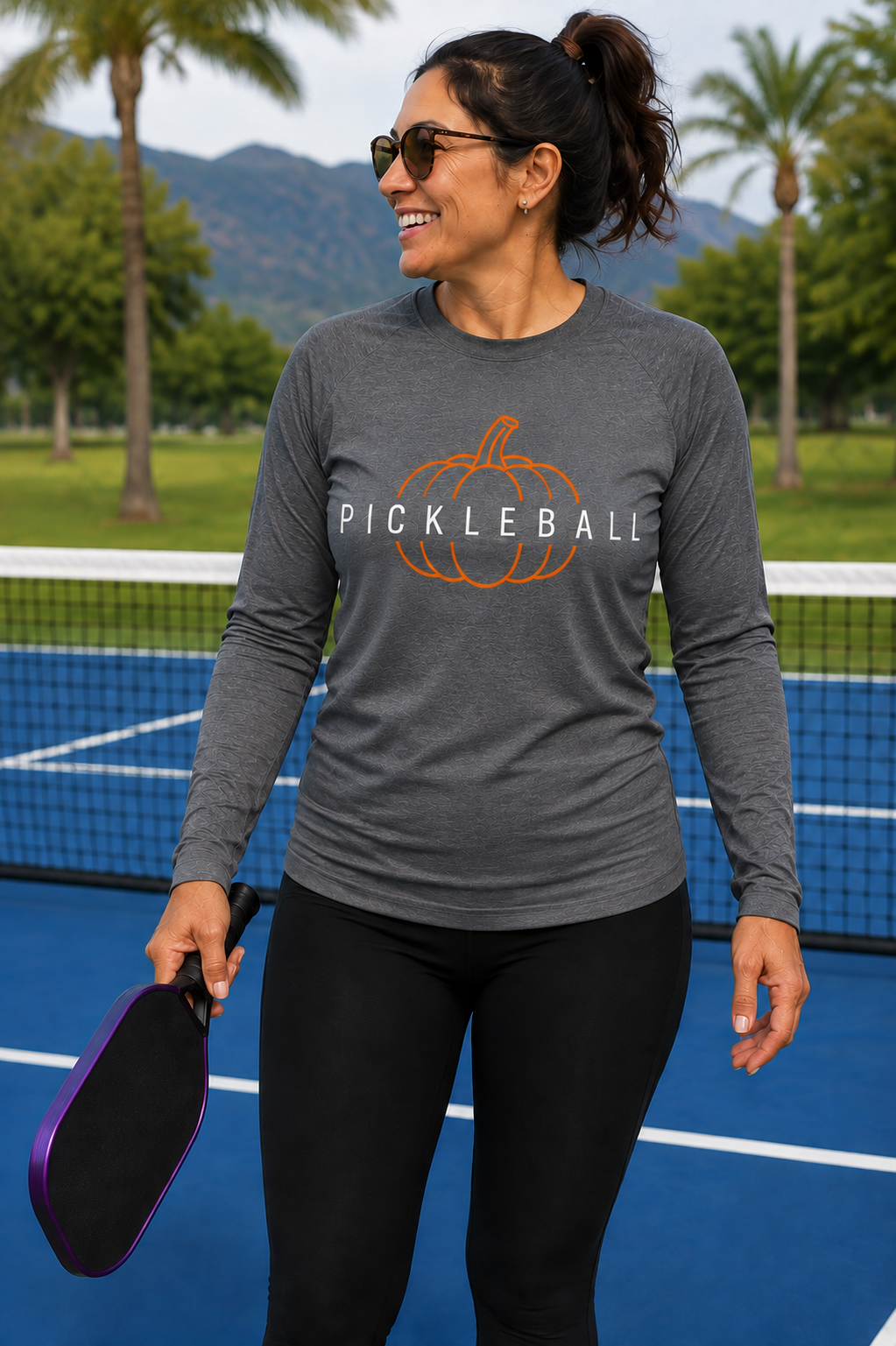 "Woman smiling on an outdoor pickleball court wearing a medium gray 'Pickleball' pumpkin graphic long-sleeve moisture-wicking shirt, holding a purple paddle with palm trees and mountains in the background"