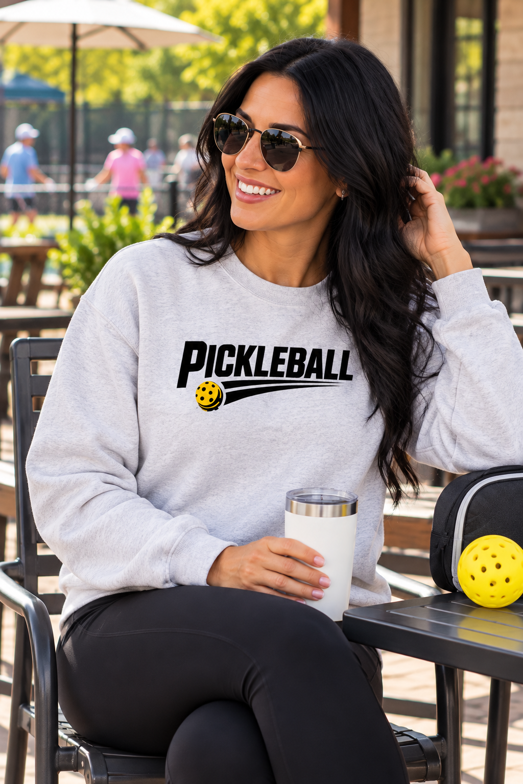 Woman wearing a light gray sweatshirt with black “PICKLEBALL” lettering and a yellow ball speed-streak graphic, seated at an outdoor table near pickleball courts.
