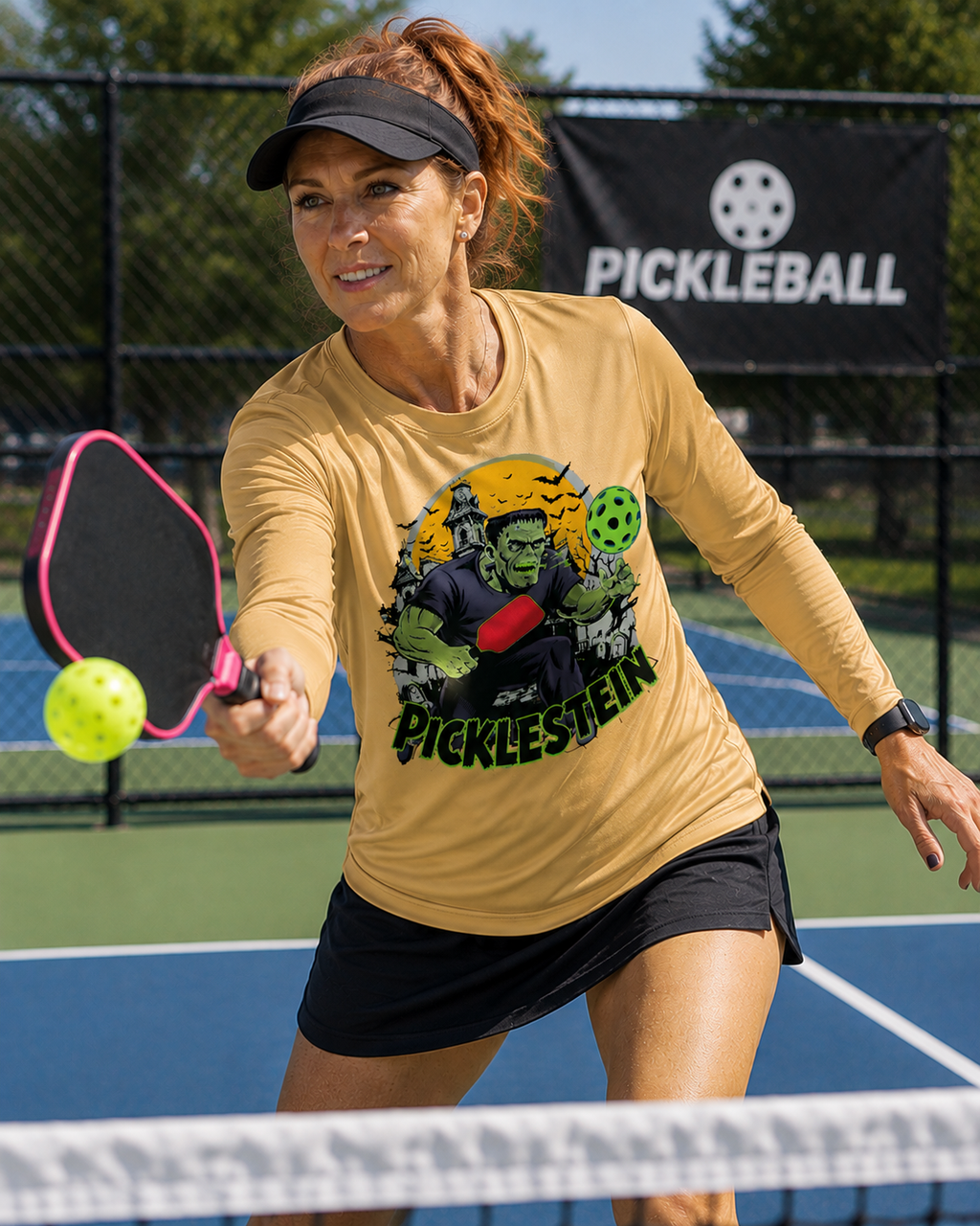 "Woman playing pickleball on a blue court wearing a sport vegas gold long-sleeve 'Picklestein' Halloween graphic tee featuring a Frankenstein monster playing pickleball, mid-swing with a pink paddle reaching for a yellow ball, with a Pickleball banner in the background."