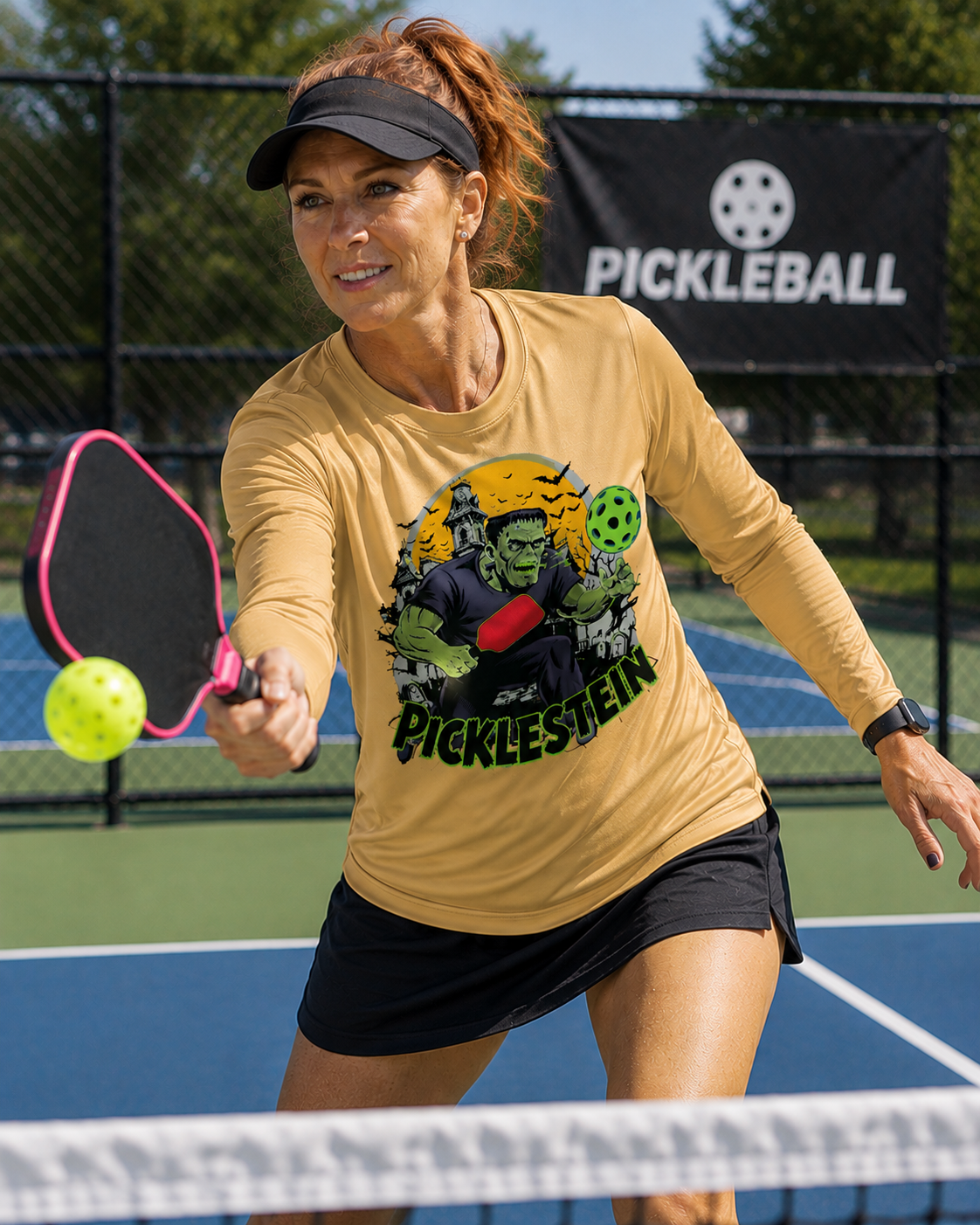"Woman playing pickleball on a blue court wearing a sport vegas gold long-sleeve 'Picklestein' Halloween graphic tee featuring a Frankenstein monster playing pickleball, mid-swing with a pink paddle reaching for a yellow ball, with a Pickleball banner in the background."