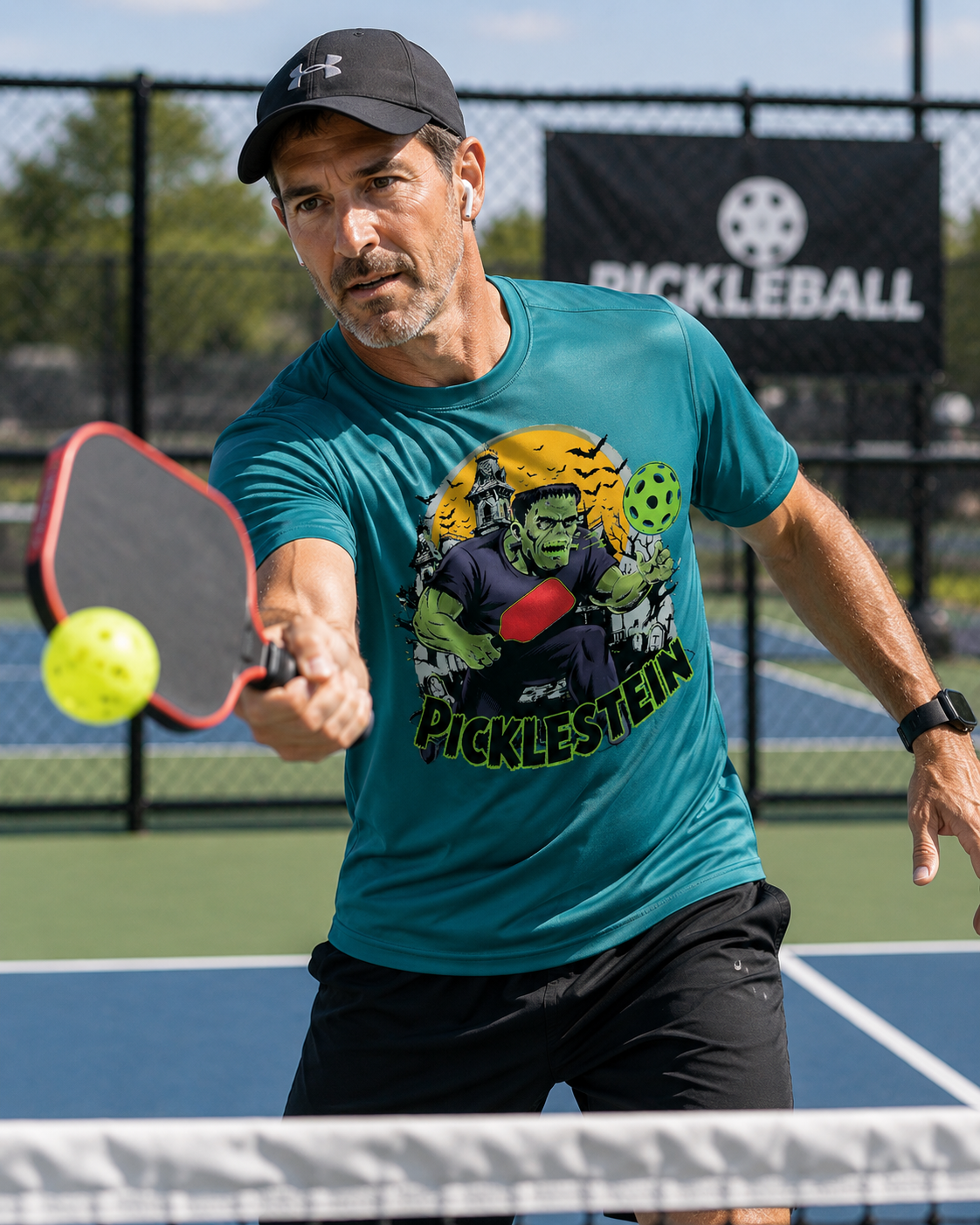 "Man playing pickleball on a blue court wearing a teal 'Picklestein' Halloween graphic tee featuring a Frankenstein monster playing pickleball, mid-swing with a red paddle reaching for a yellow ball, with a Pickleball banner in the background."