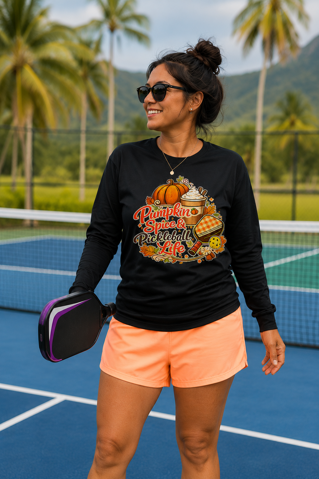 "Woman smiling on a tropical outdoor pickleball court wearing a black long-sleeve sweatshirt with 'Pumpkin Spice & Pickleball Life' fall-themed graphic design, holding a paddle with palm trees and mountains in the background"