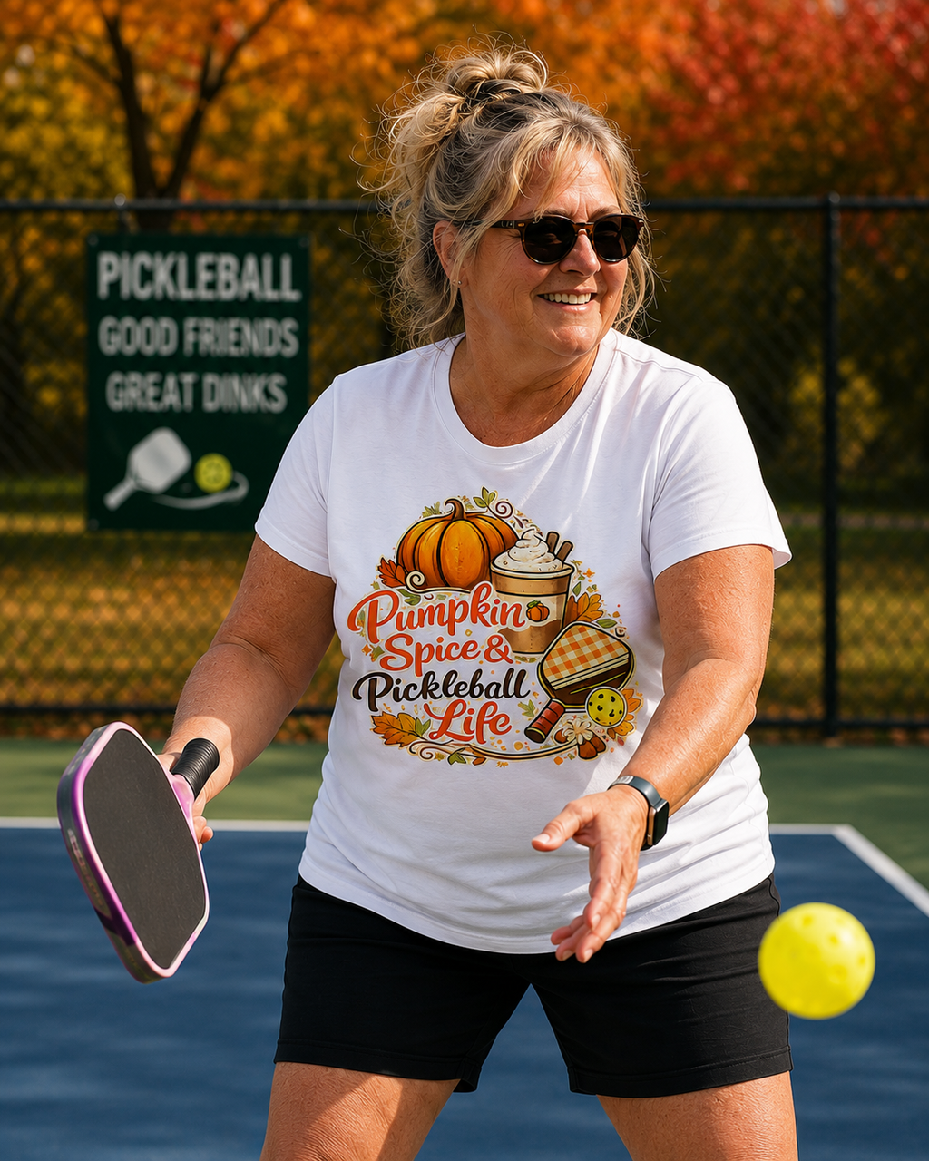 "Woman playing pickleball on an outdoor court wearing a white t-shirt with 'Pumpkin Spice & Pickleball Life' fall-themed graphic design, featuring autumn foliage in the background"