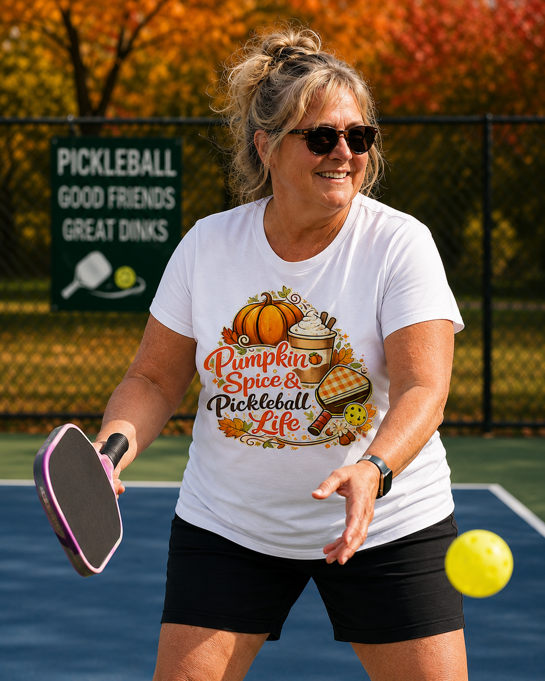"Woman playing pickleball on an outdoor court wearing a white t-shirt with 'Pumpkin Spice & Pickleball Life' fall-themed graphic design, featuring autumn foliage in the background"