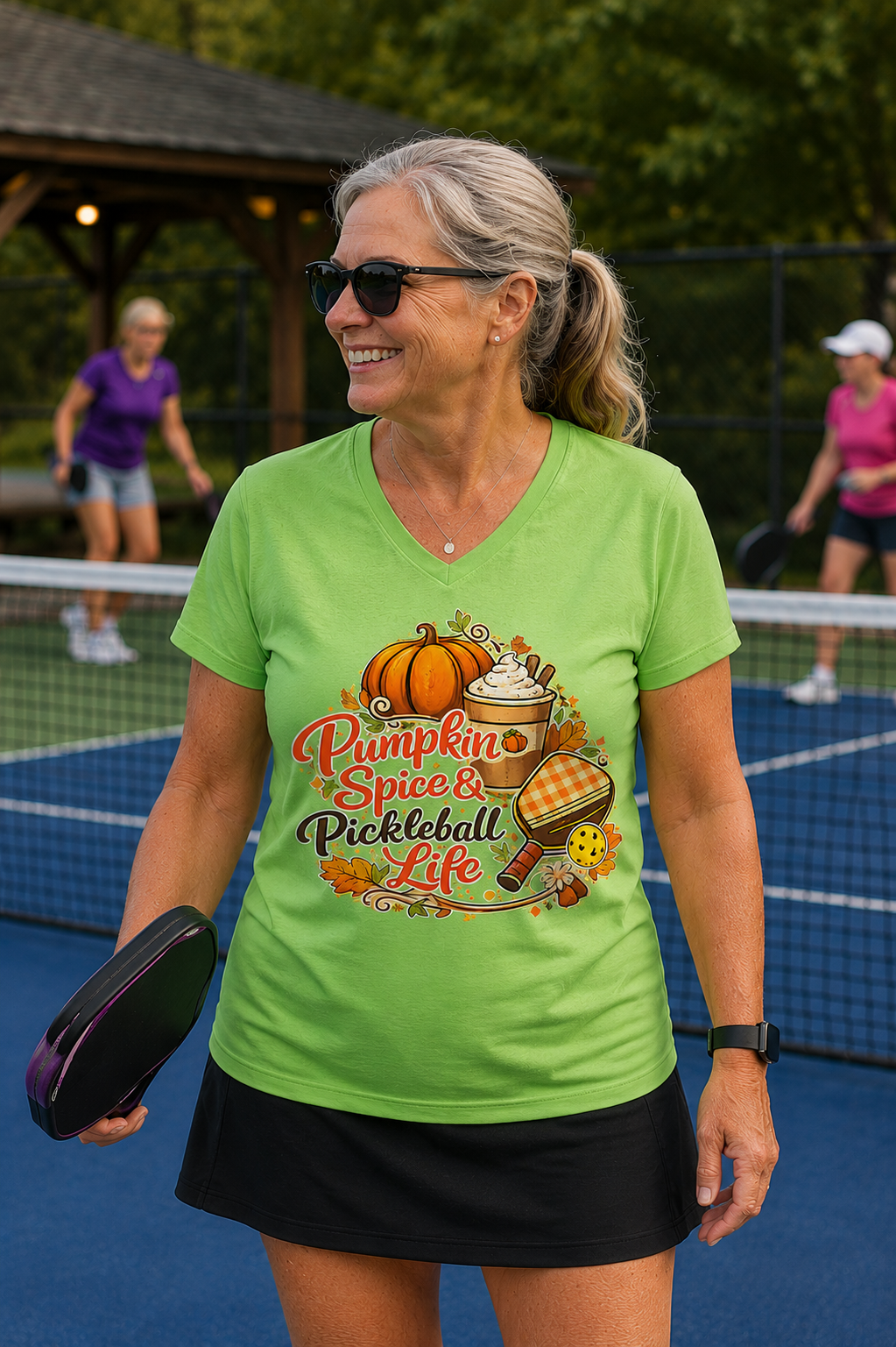 "Woman smiling on an outdoor pickleball court wearing a lime green v-neck t-shirt with 'Pumpkin Spice & Pickleball Life' fall-themed graphic design, holding a paddle with other players in the background"