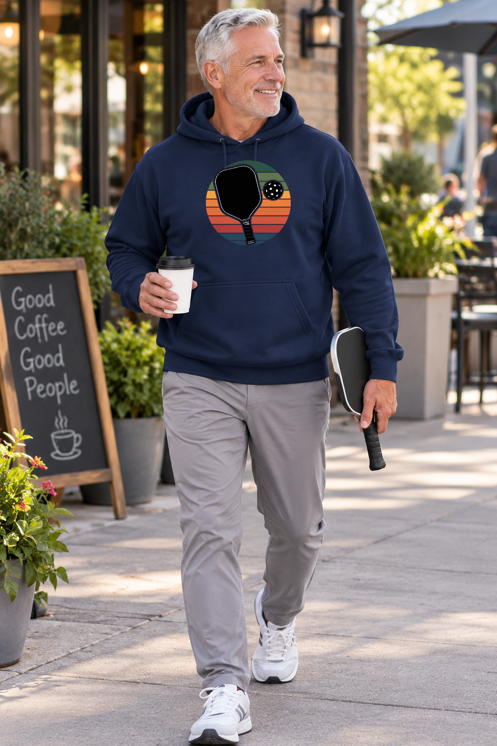 Man wearing a navy hoodie with a retro sunset pickleball paddle graphic, holding coffee and a paddle while walking outside a café.