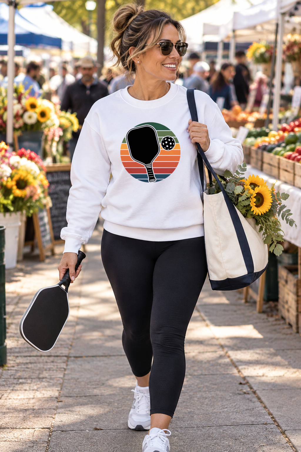Woman wearing a white sweatshirt with a retro striped pickleball paddle graphic at a farmers market, holding a paddle and carrying a tote of sunflowers.
