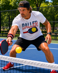 Load image into Gallery viewer, "Man playing pickleball outdoors wearing a white USA Baller patriotic graphic t-shirt, lunging low to return a shot at the net on a blue court"