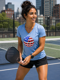 Load image into Gallery viewer, "Woman smiling on an outdoor urban pickleball court wearing a light blue v-neck t-shirt with a red, white, and blue patriotic pickleball graphic, holding a black paddle"