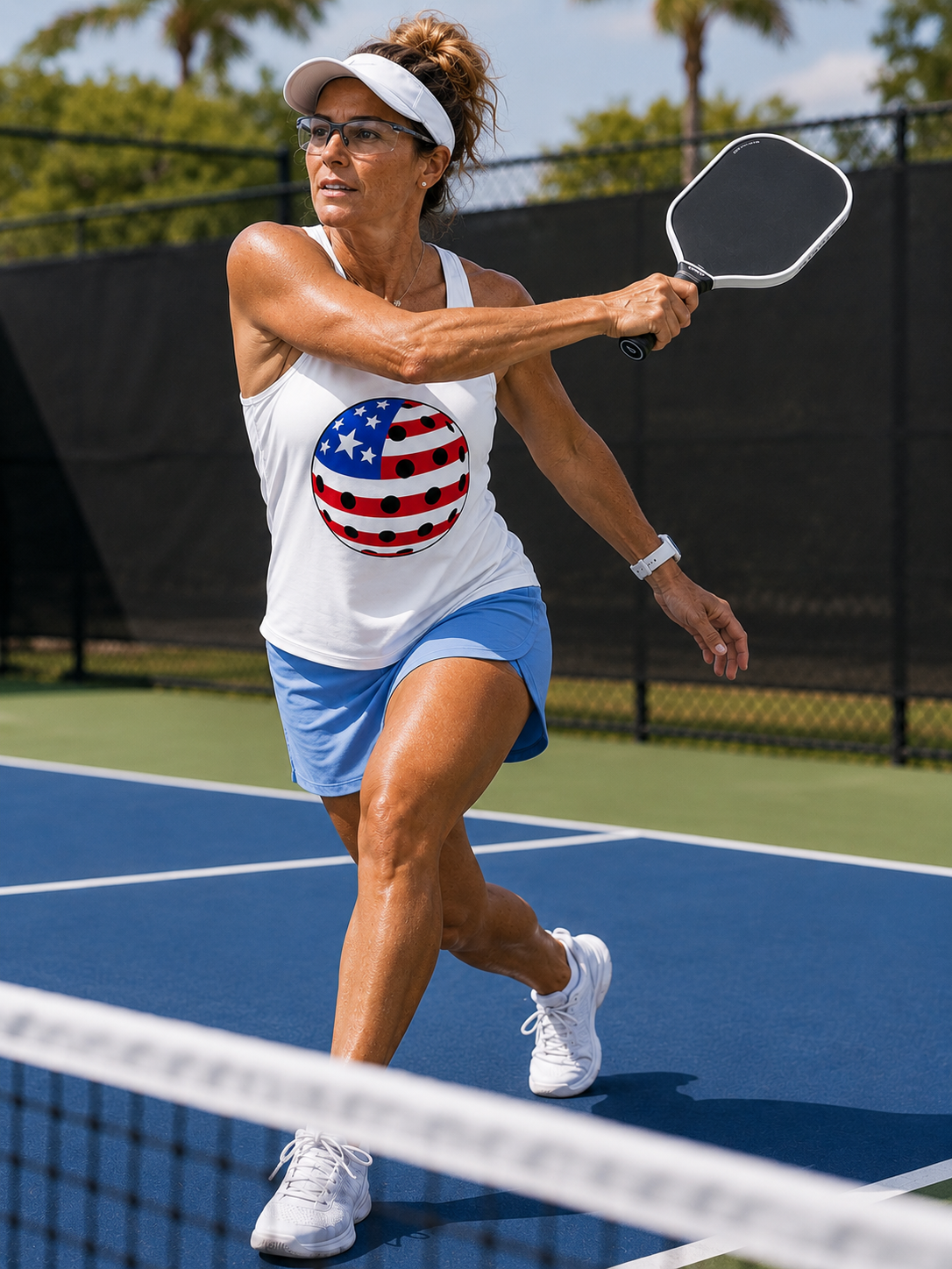 "Woman mid-swing on a pickleball court wearing a white tank top with a red, white, and blue patriotic pickleball graphic, paired with a light blue skort and white visor"
