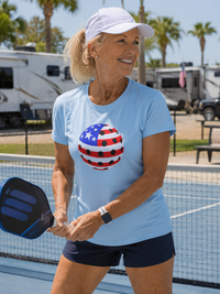 Load image into Gallery viewer, "Woman smiling on a pickleball court wearing a light blue t-shirt with a red, white, and blue patriotic pickleball graphic, holding a paddle". RV Park and palm trees.