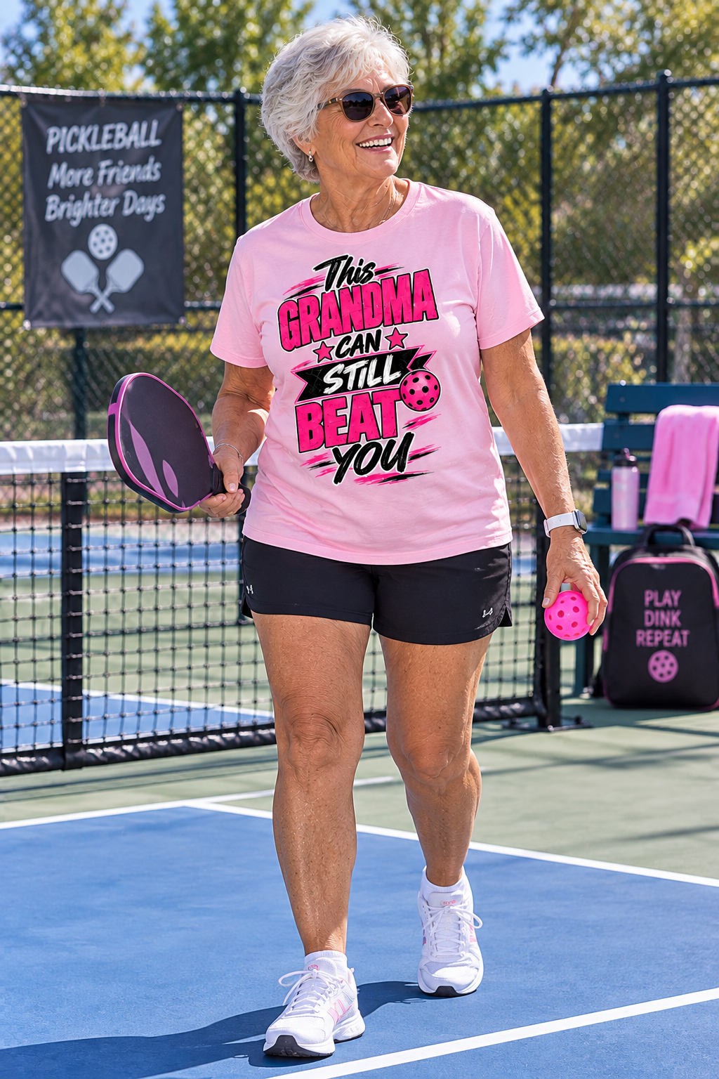 "Smiling older woman on a pickleball court holding a pink-and-black paddle and pink pickleball, wearing a light pink T-shirt printed with 'This Grandma Can Still Beat You' in pink and black lettering, paired with black athletic shorts and white sneakers."