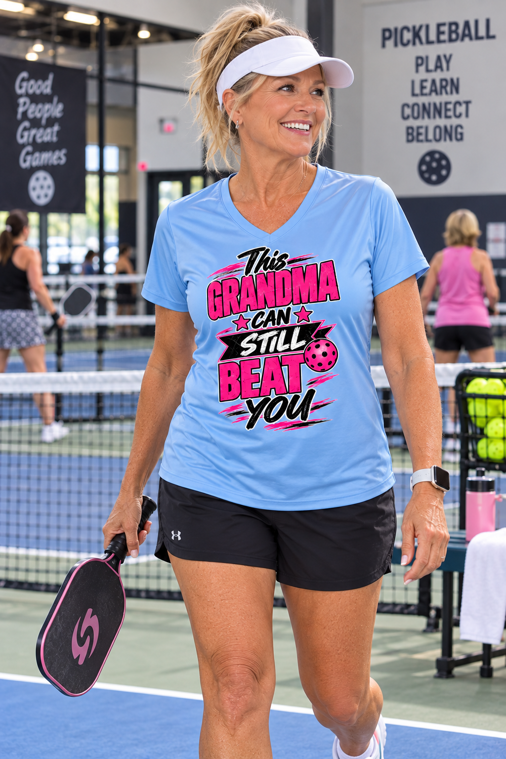 Woman wearing a light blue “This Grandma Can Still Beat You” pickleball T-shirt and holding a paddle on an indoor court.⠋