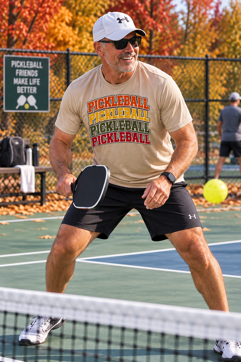 "Man playing pickleball on an outdoor court wearing a desert khaki Men's Vintage Varsity Stacked Pickleball moisture wicking graphic tee with fall foliage in the background"