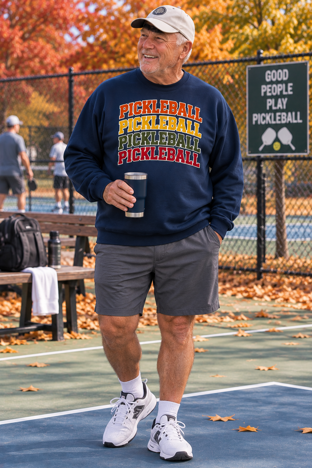 "Man standing on a pickleball court holding a tumbler wearing a navy Men's Vintage Varsity Stacked Pickleball crewneck sweatshirt with fall foliage in the background"
