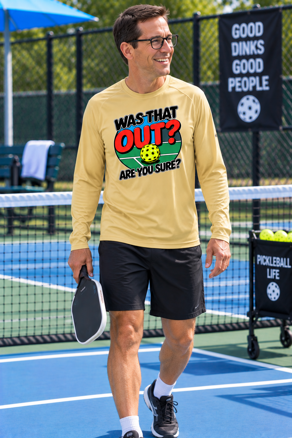 A man walking on a pickleball court wearing a beige long-sleeve t-shirt with a colorful graphic that reads 'WAS THAT OUT? ARE YOU SURE?' and black pickleball shorts.