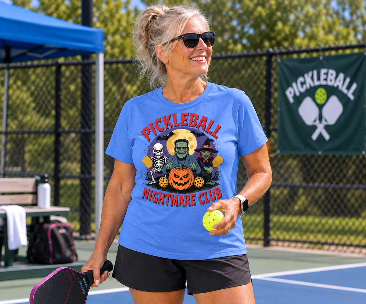 Woman wearing a blue “Pickleball Nightmare Club” Halloween T-shirt while holding a pickleball and paddle on a pickleball court.