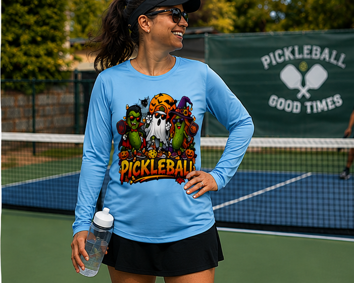 "Woman smiling on a pickleball court wearing a light blue long-sleeve Halloween-themed pickleball graphic tee and black skort, holding a water bottle, with a 'Pickleball Good Times' banner in the background."
