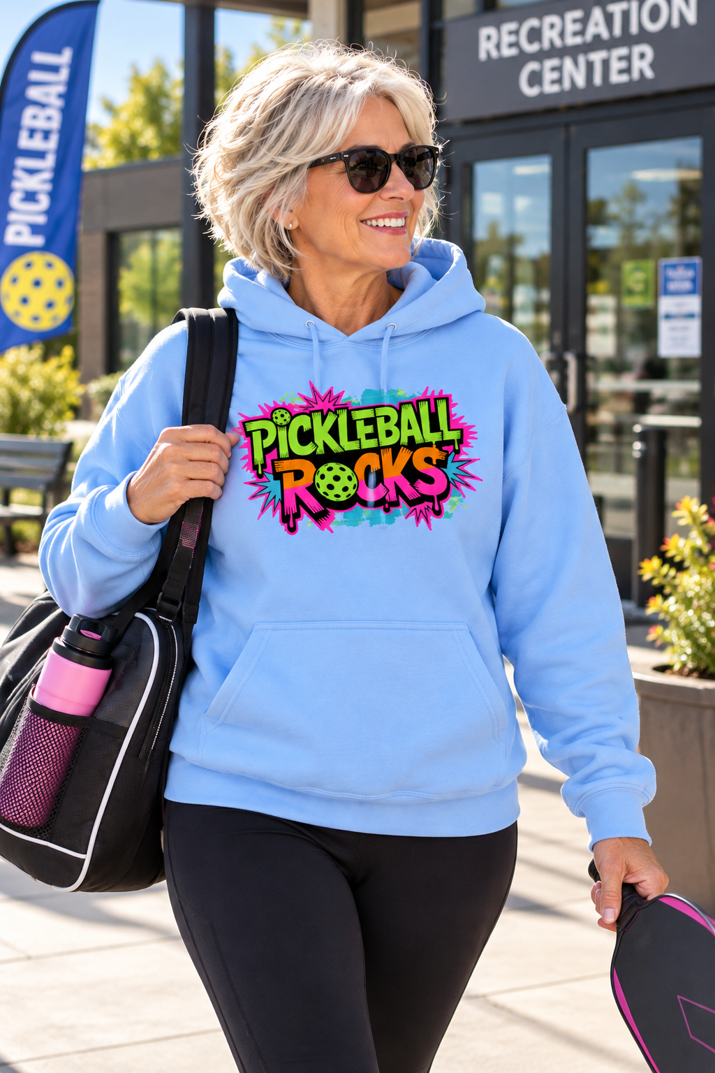 Woman wearing a light blue hoodie with a colorful retro “Pickleball Rocks” graphic, carrying a paddle and sports bag outside a recreation center.