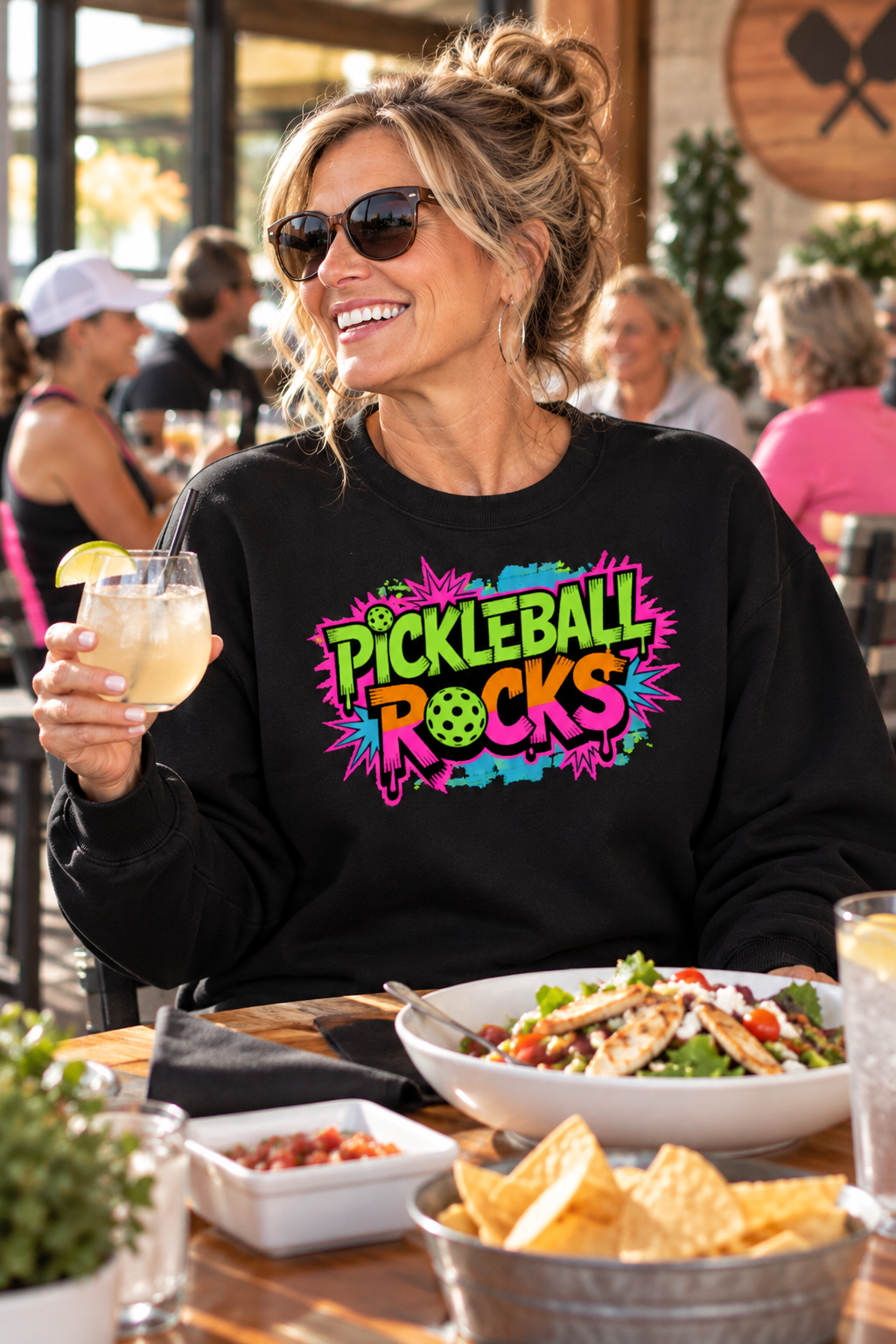 Woman wearing sunglasses and a black sweatshirt with a colorful retro “Pickleball Rocks” graphic, holding a drink at an outdoor restaurant table.